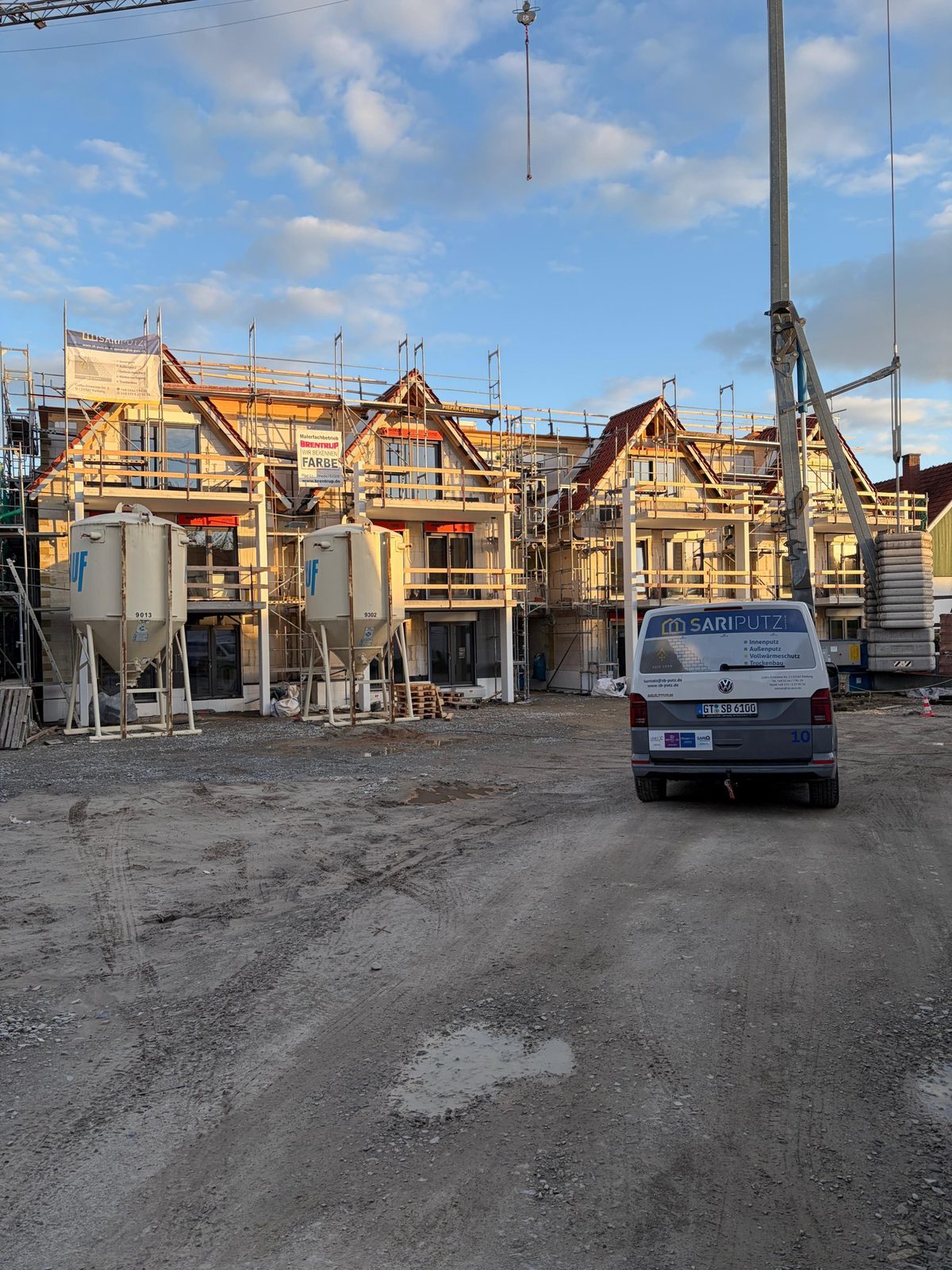 Neubau von Mehrfamilienhäusern im Rohbau mit Gerüst und SARI PUTZ-Banner, davor zwei Knauf-Putzsilos, ein Baukran und ein SARI PUTZ-Transporter